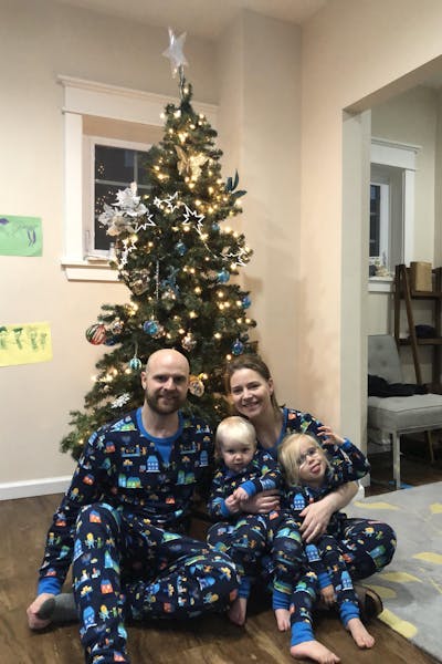 For people in pyjamas sitting in front of a christmas tree. Two are adults, two are children. The floor is wooden and the walls are beige. They are smiling.